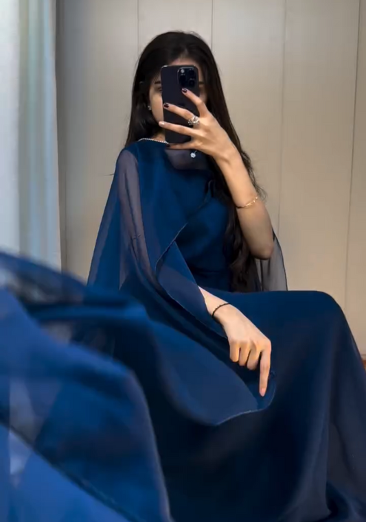 A woman stands in front of a neutral background wearing a navy blue cape dress paired with a silver mini handbag, posing with her phone covering her face.